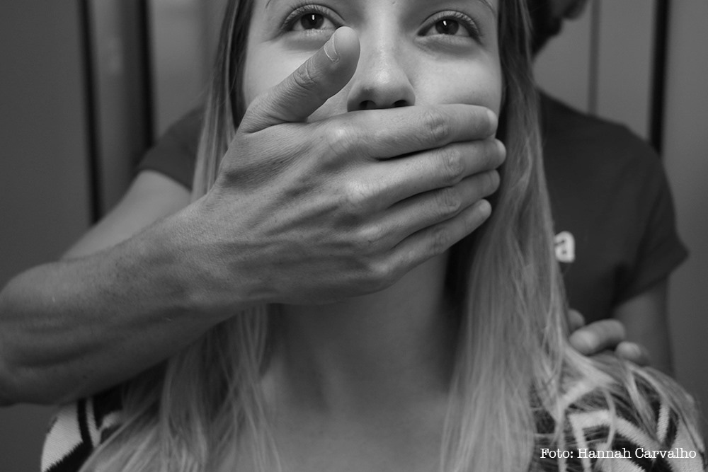 imagem de uma mulher branca de cabelos loiros olhando para o alto, enquanto um homem do qual só vemos o tronco, está tapando a boca dela.