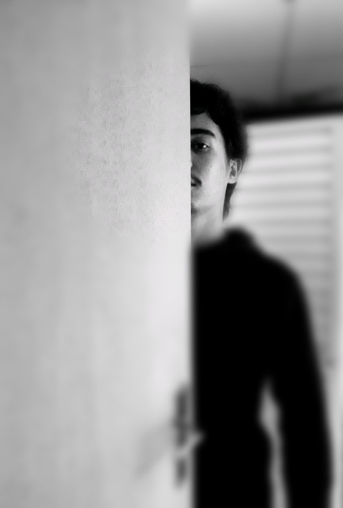 imagem de um menino atrás de uma parede, só se vê metade de seu corpo, ele é branco de cabelos pretos lisos e curtos, veste uma blusa de frio preta e tem um olhar impassível.