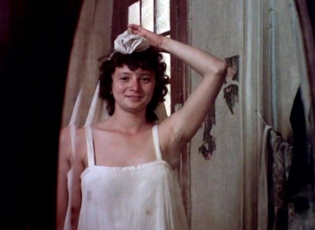 Mulher branca de cabelos escuros, curtos e ondulados. Ela está se olhando sorrindo no espelho, usa um vestido branco, e segura com um braço um tecido branco no topo da cabeça.