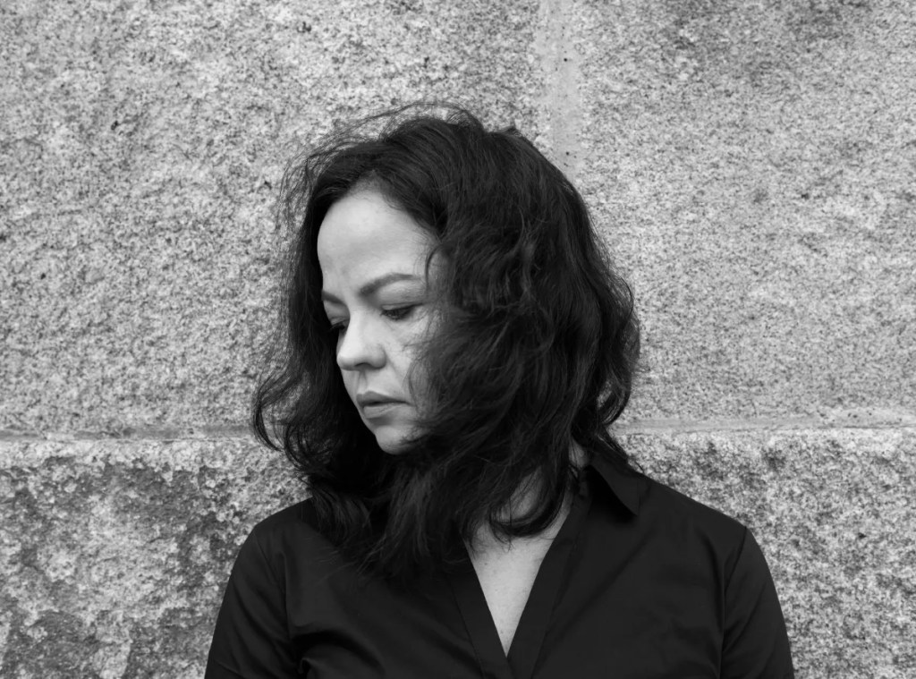 Foto em preto e branco em plano médio de uma mulher de cabelos escuros, de perfil, olhando para baixo. Ela veste camisa preta com leve decote. 