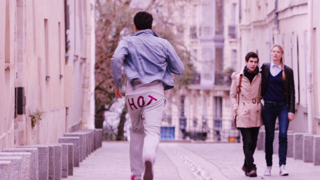 A imagem mostra um homem correndo com uma calça escrits "hot" (quente) na parte de trás. No outro lado da rua, há um casal com uma mulher abraçando o parceiro de forma protetora. 