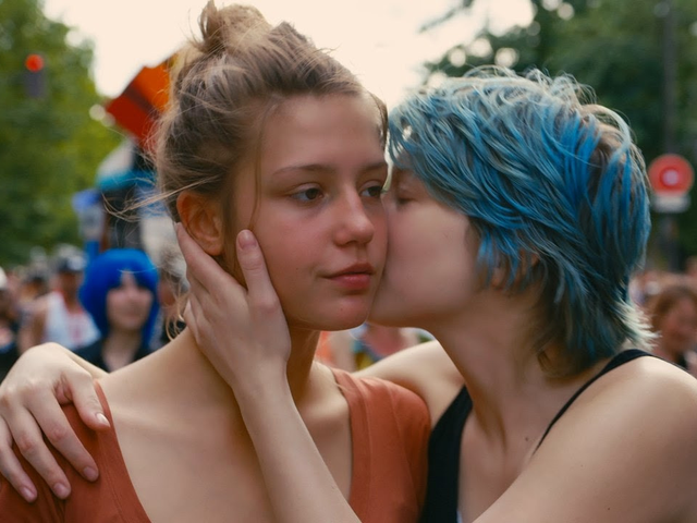 Na imagem, Emma, uma das protagonistas, beija a bochecha de Adèle. Elas estão em primeiro plano e em um ambiente público, que parece ser bastante colorido.