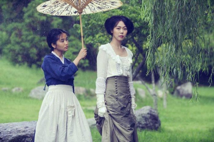 Na foto, Sook-Hee segura um guarda chuva para Hideko, que olha diretamente para a câmera. As personagens estão em um cenário arbóreo, como uma floresta