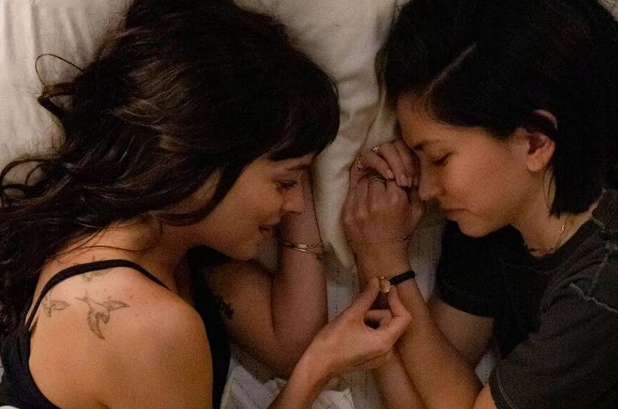 na imagem aparecem duas mulheres deitadas em uma cama, uma de frente para outra. a da esquerda é branca e tem cabelo liso com franja. e a outra é de origem asiática e tem cabelo preto curto.