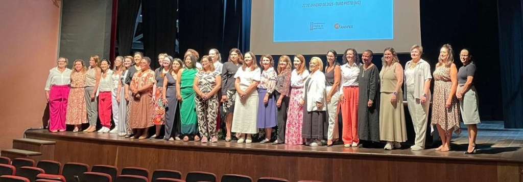 Na imagem, palestrantes e integrantes da Andifes se posicionam na parte frontal do palco de apresentação. Elas utilizam roupas diversas e coloridas.  