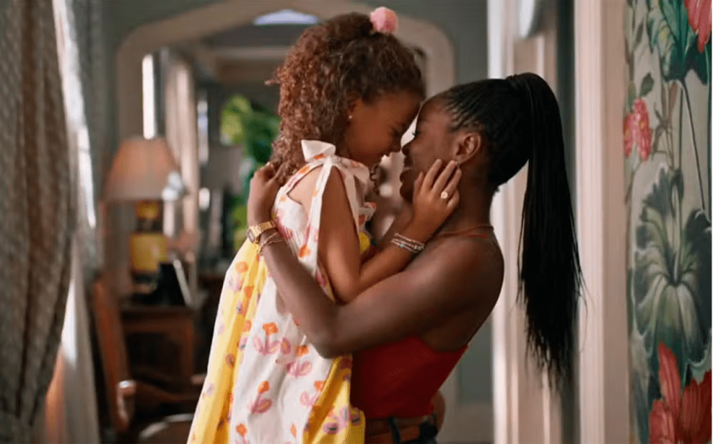 Uma mulher negra abraça no colo uma menina negra, com alegria em um ambiente familiar. As duas se olham, e a menina tem a mão no rosto da mulher.