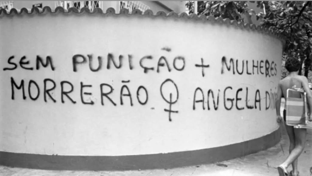 Uma imagem em preto e branco mostra um protesto grafite com mensagens em português dizendo: “Sem punição, mais mulheres morrerão”. A seguir, está escrito o nome Angela Diniz