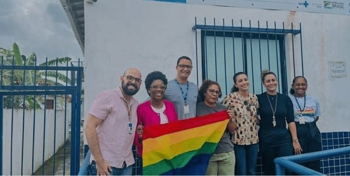 Grupo de pessoas reunidos em frente a prédio, segurando a bandeira do arco-íris. 