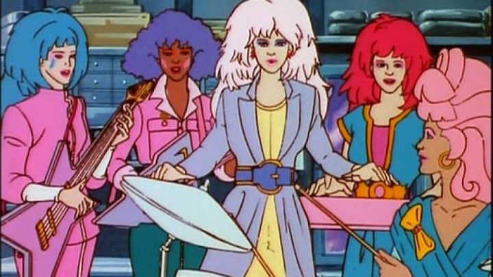 Imagem de desenho animado de cinco mulheres com cabelos estilo anos 80 em cores chamativas rosa, azul, lilás, vermelho e pink. Elas tocam instrumentos de rock.