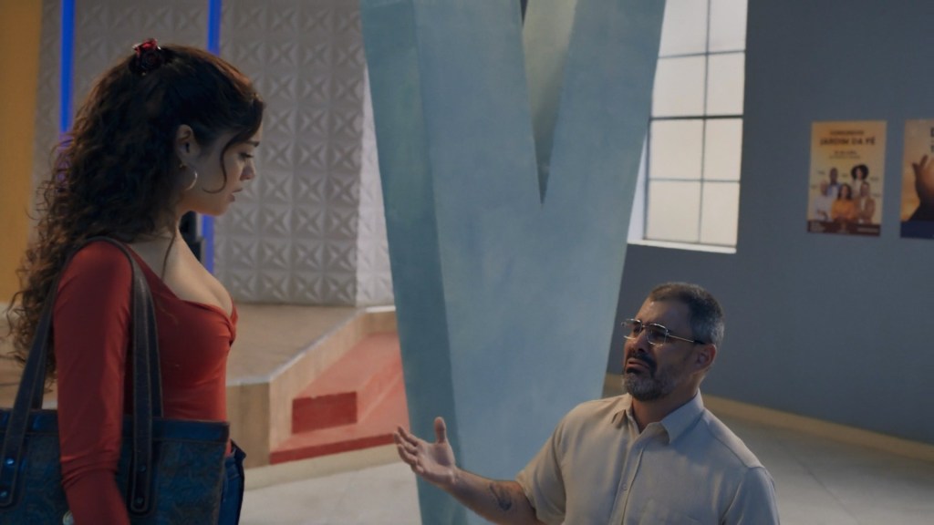 Homem ajoelhado, chorando, de mãos erguidas, é olhado de cima por mulher de blusa vermelha. 