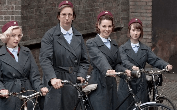 Quatro mulheres com sobretudo cinza, blusas azuis e chapéus vermelhos estão ao lado das suas bicicletas antigas.