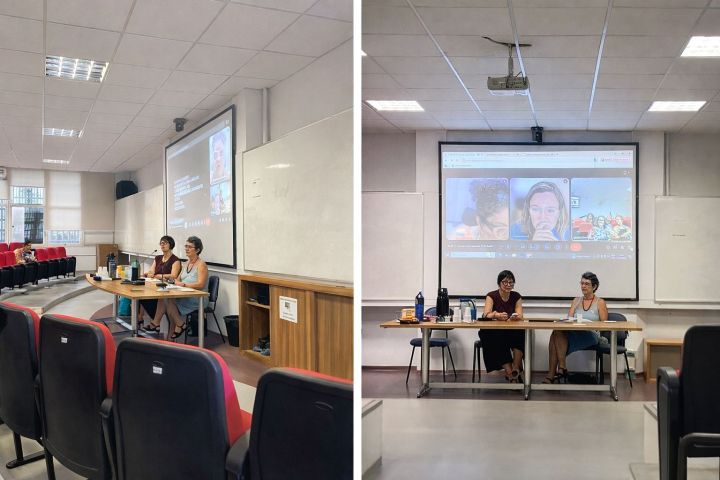 Duas mulheres sentadas em cadeiras em um auditório, à frente de uma tela de projetor.