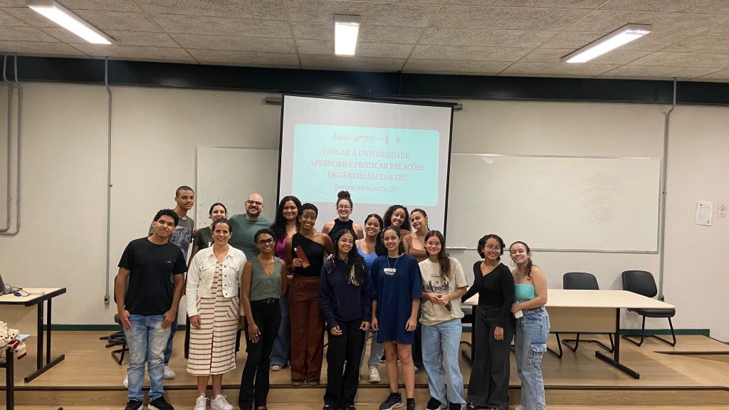 Grupo de 17 pessoas posando para foto em uma sala de aula. Ao fundo, há um projetor exibindo um slide com o título “Chegar à universidade: relações de gênero saudáveis”. As pessoas estão organizadas em duas fileiras, sorrindo, com roupas casuais. A sala tem quadro branco, mesas, cadeiras e iluminação fluorescente no teto.