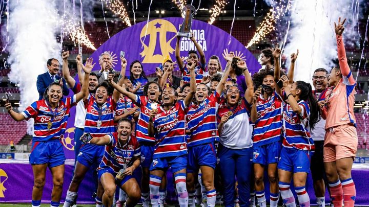 A imagem captura um momento de pura euforia e triunfo do time feminino do Fortaleza Esporte Clube. No centro do gramado, sob a luz dos refletores e o brilho de fogos de artifício que saltam ao fundo, as jogadoras celebram a conquista de um título importante. Elas vestem o tradicional uniforme tricolor, com listras horizontais em azul, vermelho e branco, e muitas trazem medalhas de ouro penduradas no pescoço.

O ponto focal da cena é o troféu prateado, erguido bem alto por uma das atletas, simbolizando a vitória coletiva. Ao redor dela, o grupo transborda alegria: algumas jogadoras gritam de felicidade, outras sorriem abertamente para as câmeras ou usam seus próprios celulares para registrar a eternidade do momento. Atrás do grupo, um grande painel roxo com a marca da competição emoldura a cena, enquanto a fumaça branca das celebrações pirotécnicas preenche o ar, conferindo uma atmosfera épica e vibrante à fotografia. É uma celebração que exala união, força e o orgulho de levar o troféu para casa.
